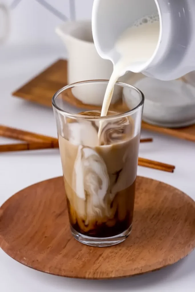 Hojicha latte being made with milk pouring into glass creating beautiful swirled patterns on wooden coaster