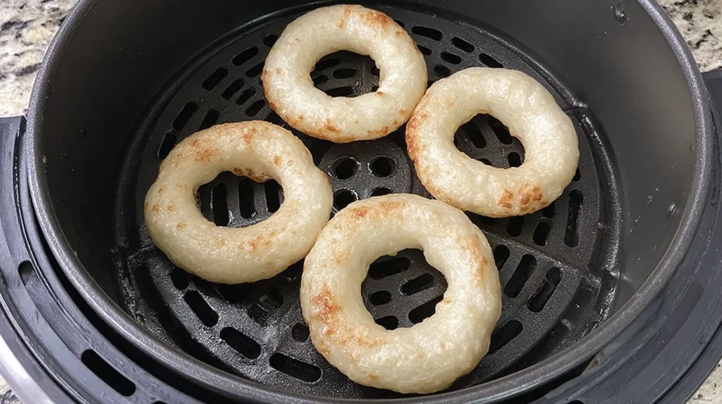Four unglazed protein donuts cooking in an air fryer basket, showing their golden-brown spots beginning to form on the pale dough surface.
